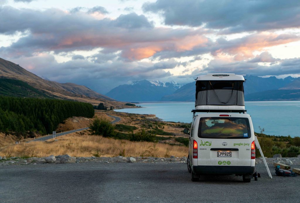 A long dusty road, mountains to the left and in the distance. We see a van parked up with the roof tent erected. Stopping for the night to take in the breathtaking views during their van life.