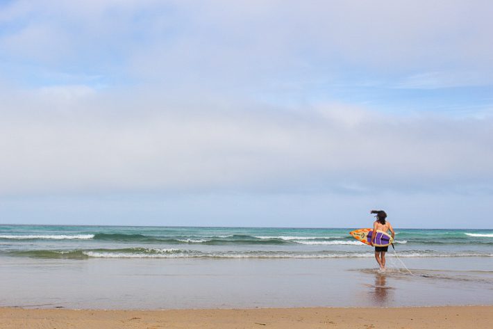 A photograph of a young man running into the ocean in Cornwall. This place is his home. He carries a surfboard and runs into the white water as the waves lap the shore. It is a sunny day and all is calm. 