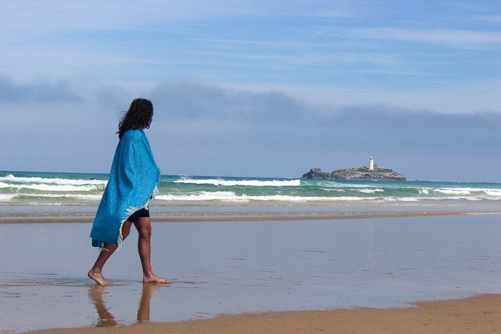 A man walks along the shoreline of a beach in Cornwall, his home. The day is sunny and the waves lap the shore. He wears a bright blue blanket as he walks. In the background is a lighthouse. 