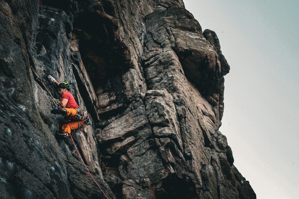 A man can be seen climbing. He is wearing bright red and orange and climbs in a harness and helmet. He is clinging on to the cliffside in Cornwall during one of his adventures. 