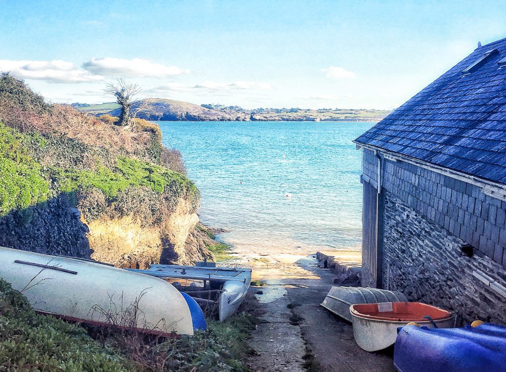 A photo of a cove in Cornwall on a hiking trail. It is a sunny day and the waters are blue. Small boats are in the foreground of the image. 