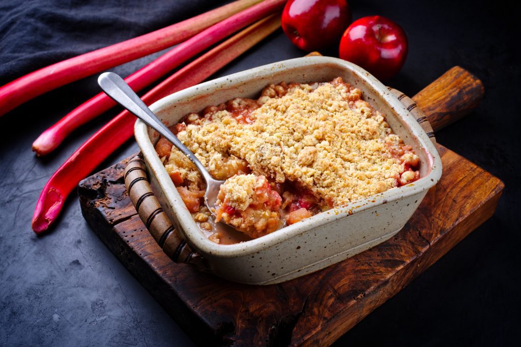 A photograph of crumble ready to eat. Surrounding this is chunks of rhubarb ready to make into crumble. 