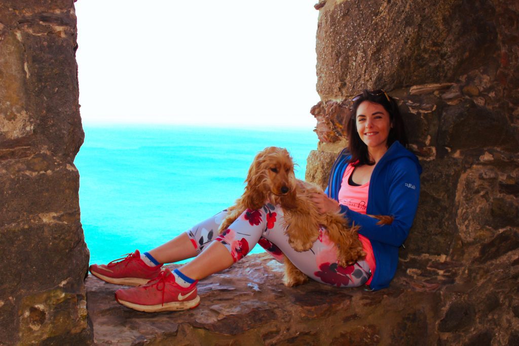 A photo of a woman and her dog sat inside a mine engine in Cornwall. They are smiling. The sea can be seen in the background. They are our for a hike. 