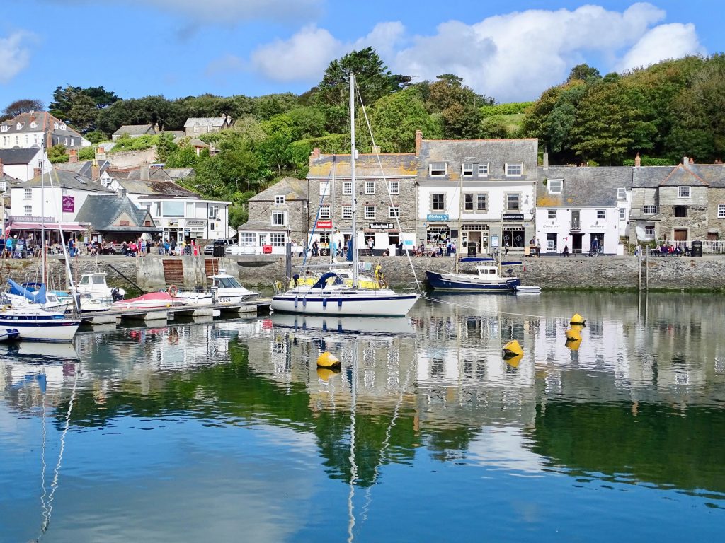 A photo of the village of Padstow in Cornwall. It is a sunny day and boats fill up the harbour. Their reflections are clear in the blue water. The pubs and shops line the background of the image. This a great place for a hike.