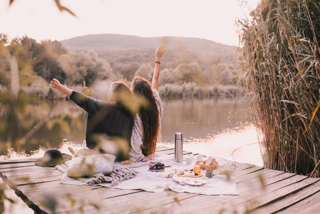 Two female friends in knitted warm sweaters having picnic near lake with autumn forest and lake on the background. Cozy fall atmosphere.