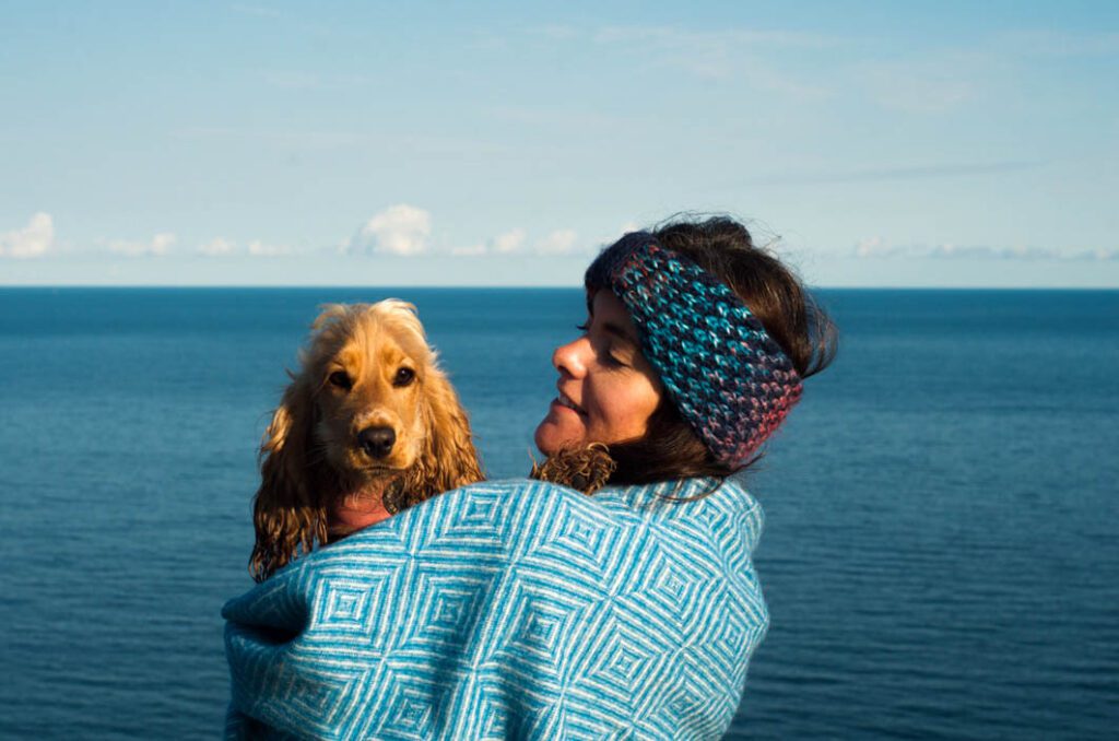 A woman and a small, fluffy dog wrapped up in a blanket looking out over the ocean. 