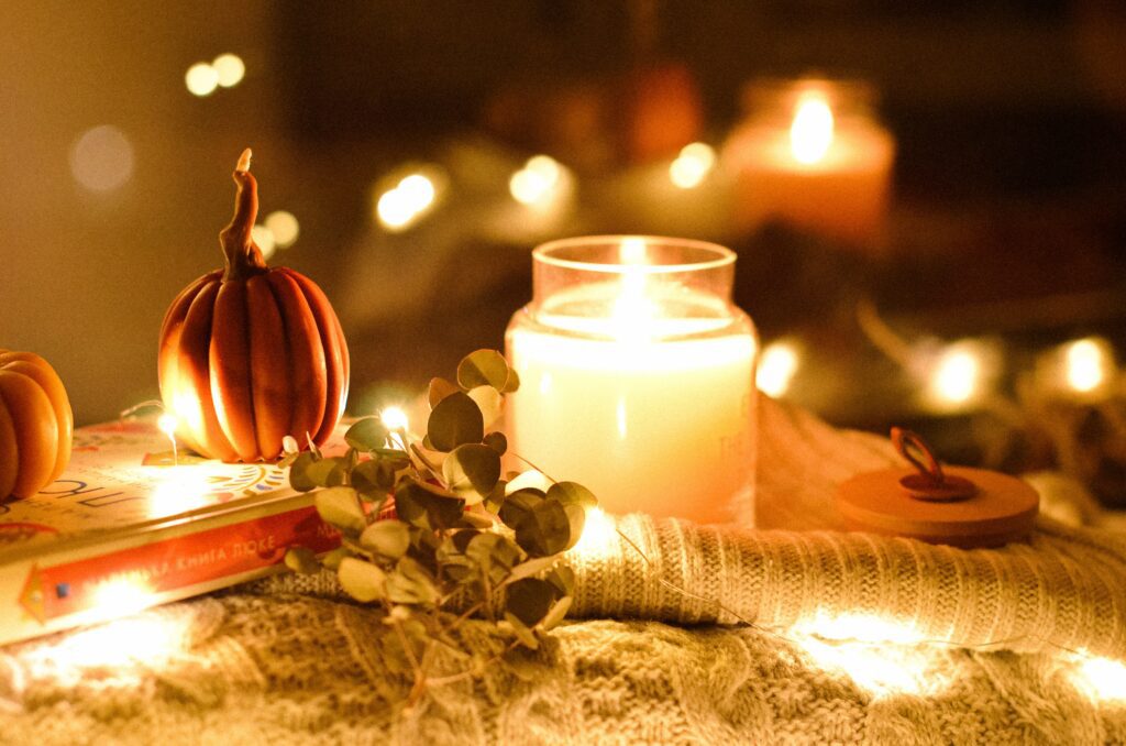 A cosy photo of a lit candle burning and lots of fairy lights. They sit atop of warm jumper and a book. 