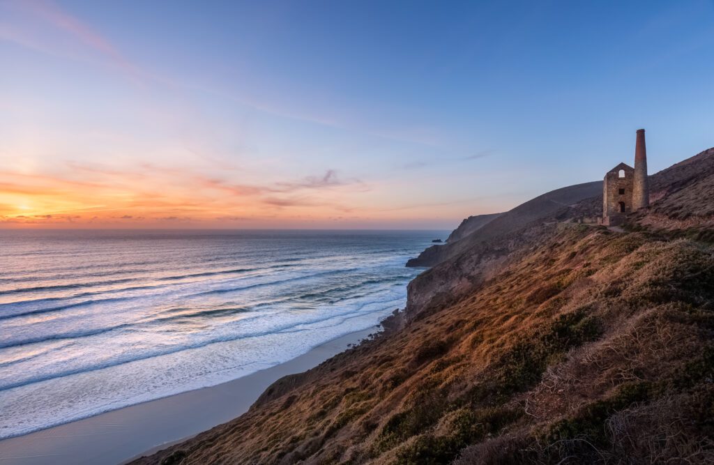 Sunset over the Wheal Coates engine house. On the left is the sea with the sun going down in an orange haze. On the right is the grass and gorse of the coast path with the engine house sitting on the path.