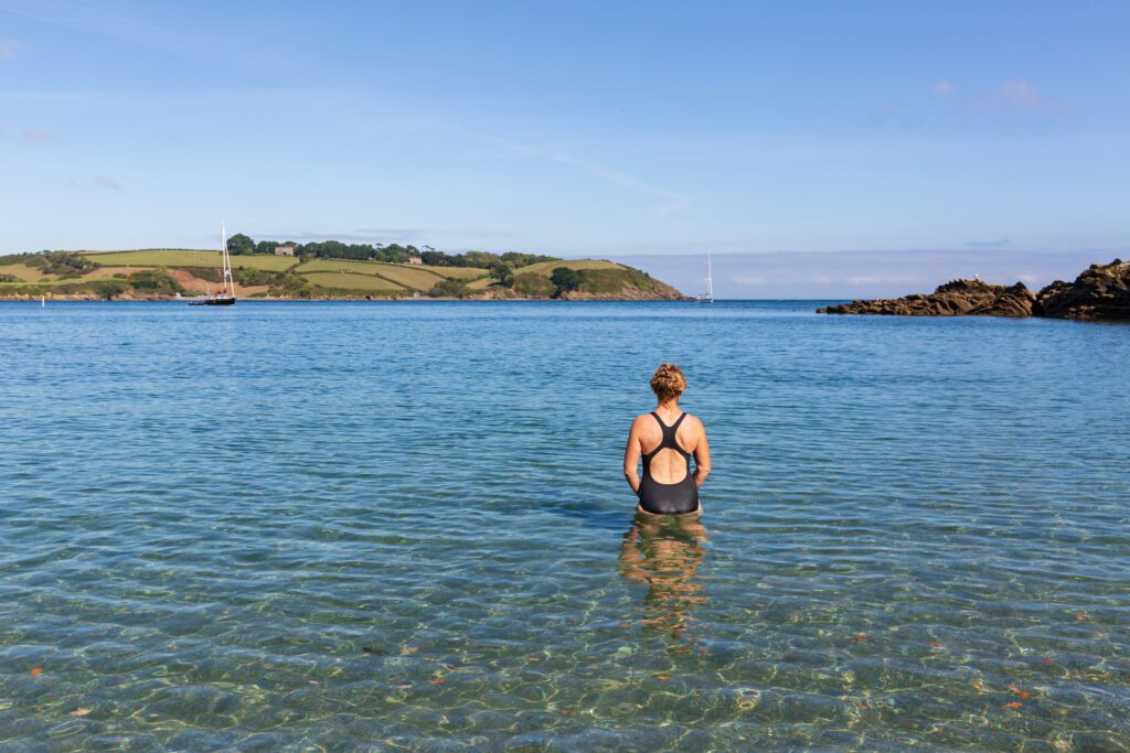 Woman wearing a swimming costume entering the cool, clear water at Bosahan Cove on the the Helford River Estuary in Cornwall, England. 