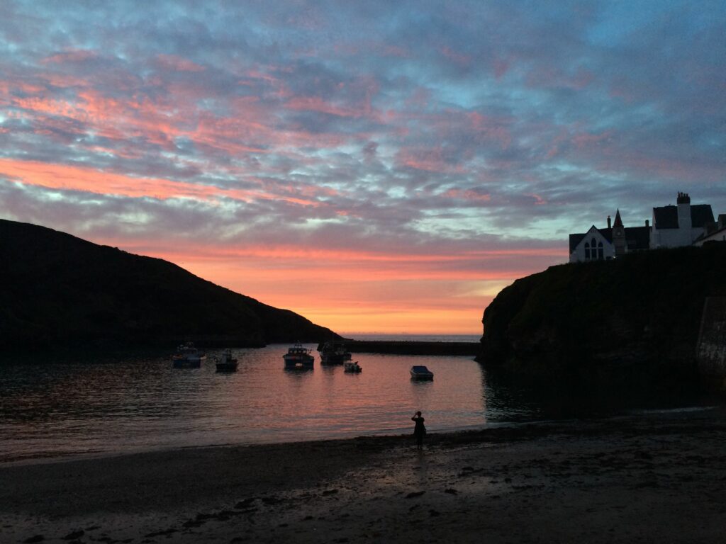 Sunset in Port Issac harbour. There are some boats gently floating in the calm waters of the harbour inside the harbour wall. The sky is orange, pink and blue with lots of broken clouds. 